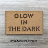 A tan woven patch with black text that reads GLOW IN THE DARK on a wooden surface.