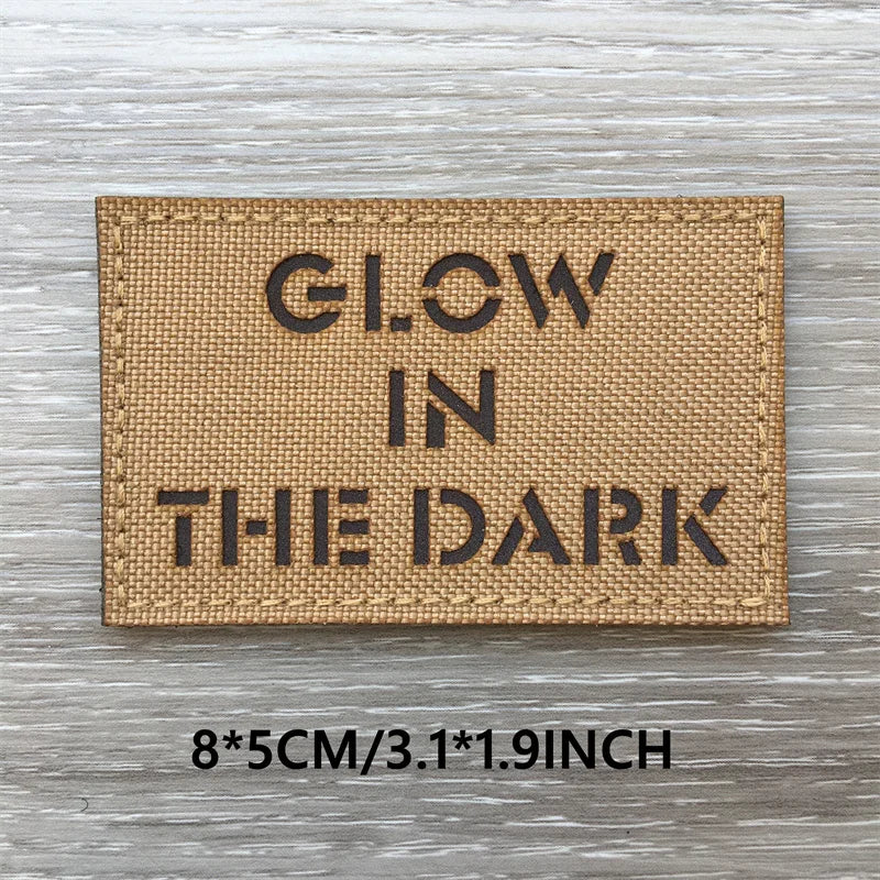 A tan woven patch with black text that reads GLOW IN THE DARK on a wooden surface.