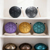 A collection of steel tongue drums in various colors including black, silver, purple, yellow, teal, and bronze displayed on w