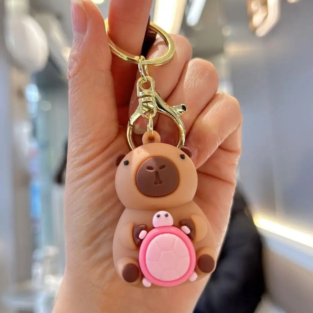 A hand holds an adorable capybara keychain charm with a gold ring and pink accents.