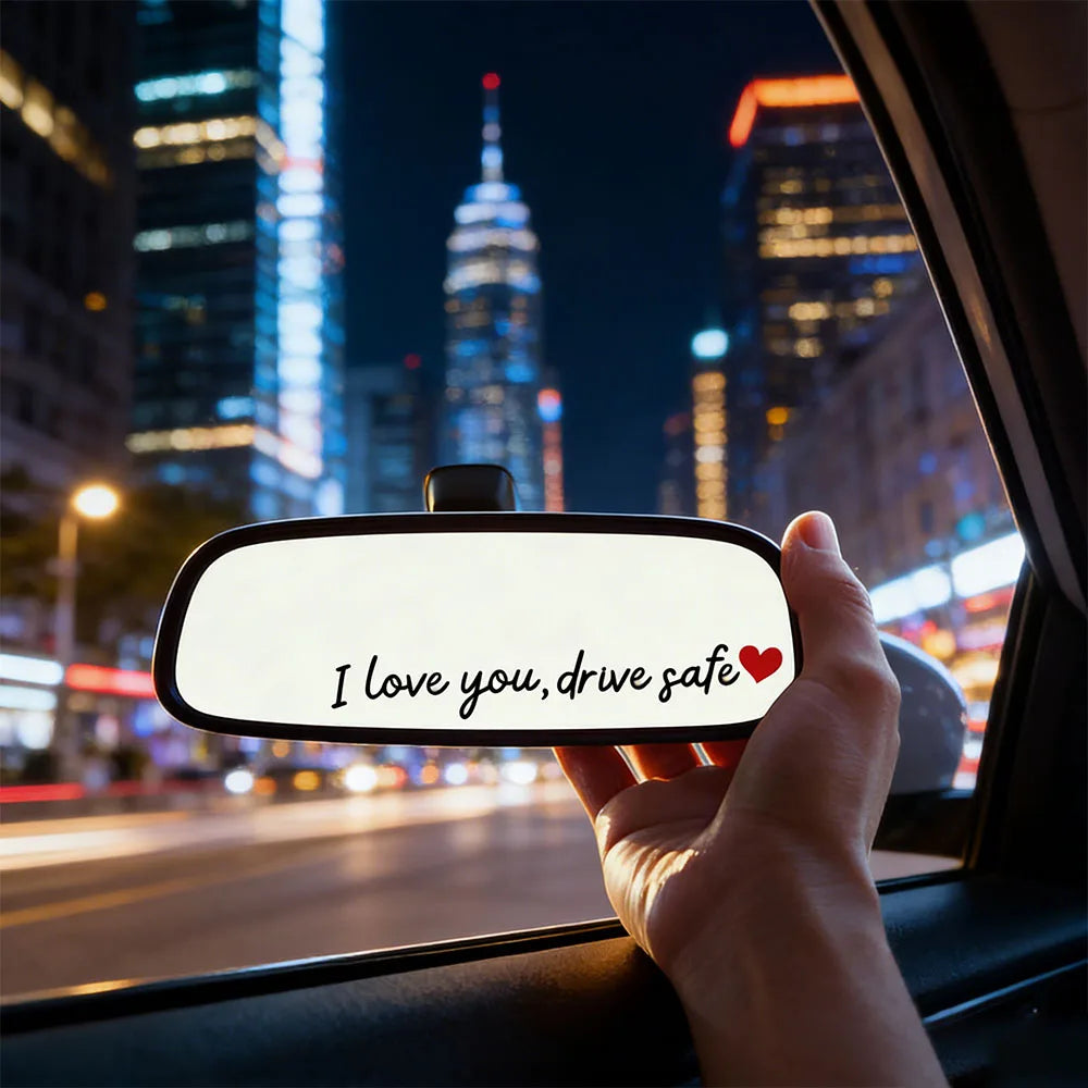 A hand holds a car mirror with the text I love you drive safe displayed on it against a city night backdrop.