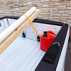 A white foam rod is tied to a wooden plank and placed inside a black truck tailgate, next to a red bottle and a black strap.