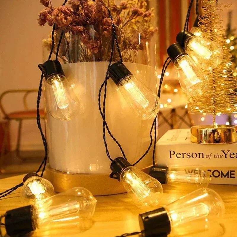 A close-up of vintage Edison bulbs strung on black wires, illuminating a white vase and warm light.