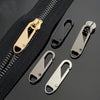 A set of eight metal zipper sliders and a gold-colored zipper pull are displayed on a black fabric background.