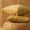 Two mustard yellow cotton linen throw pillows with tassels resting on a woven natural fiber surface.