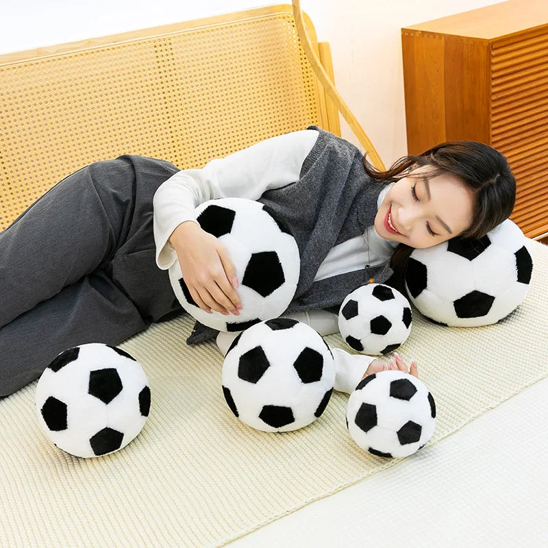 A woman is relaxing on a bed, holding and playing with a white football plush pillow.