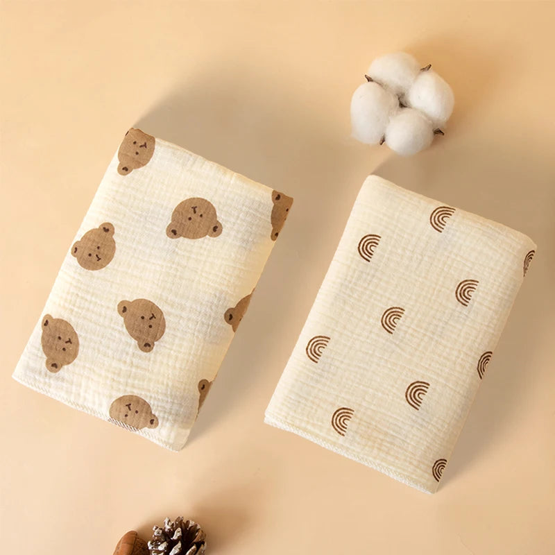 A beige soft cotton gauze baby swaddle blanket with brown bear patterns and a fluffy white pom-pom.
