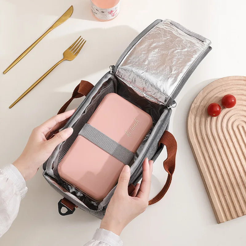 A hand is opening a pink compact thermal lunch tote bag with a silver foil lining, next to a fork and a wooden cutting board.