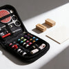 A black compact travel sewing kit is shown open, displaying various hand sewing tools and wooden items on a white surface.