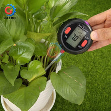 A hand holds a black soil tester device next to a potted green plant, used for measuring soil pH, moisture, and light.