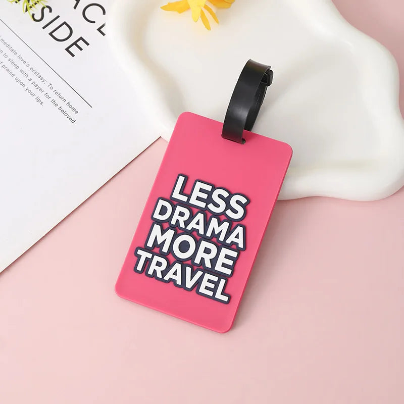 A pink silicone luggage tag with the text Less Drama More Travel is displayed on a light surface.