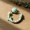 A light green pacifier clip bracelet featuring a ball, a crown charm, and the name BABY.