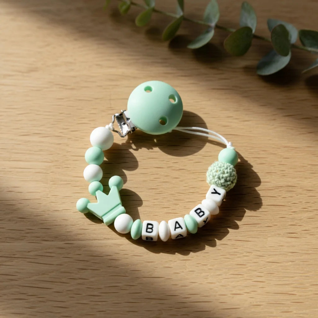 A light green pacifier clip bracelet featuring a ball, a crown charm, and the name BABY.