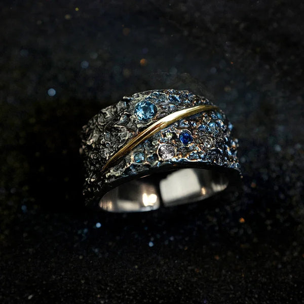 Primer plano de un anillo de plata chapada con piedras azules y banda de oro, sobre una superficie oscura y texturizada.