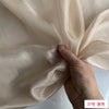 A hand is folding a piece of light beige or ivory silk chiffon fabric.