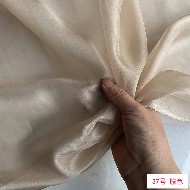 A hand is folding a piece of light beige or ivory silk chiffon fabric.