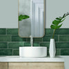 A white vessel sink is mounted on a light wood countertop against a dark green subway tile backsplash with a mirror and tropi