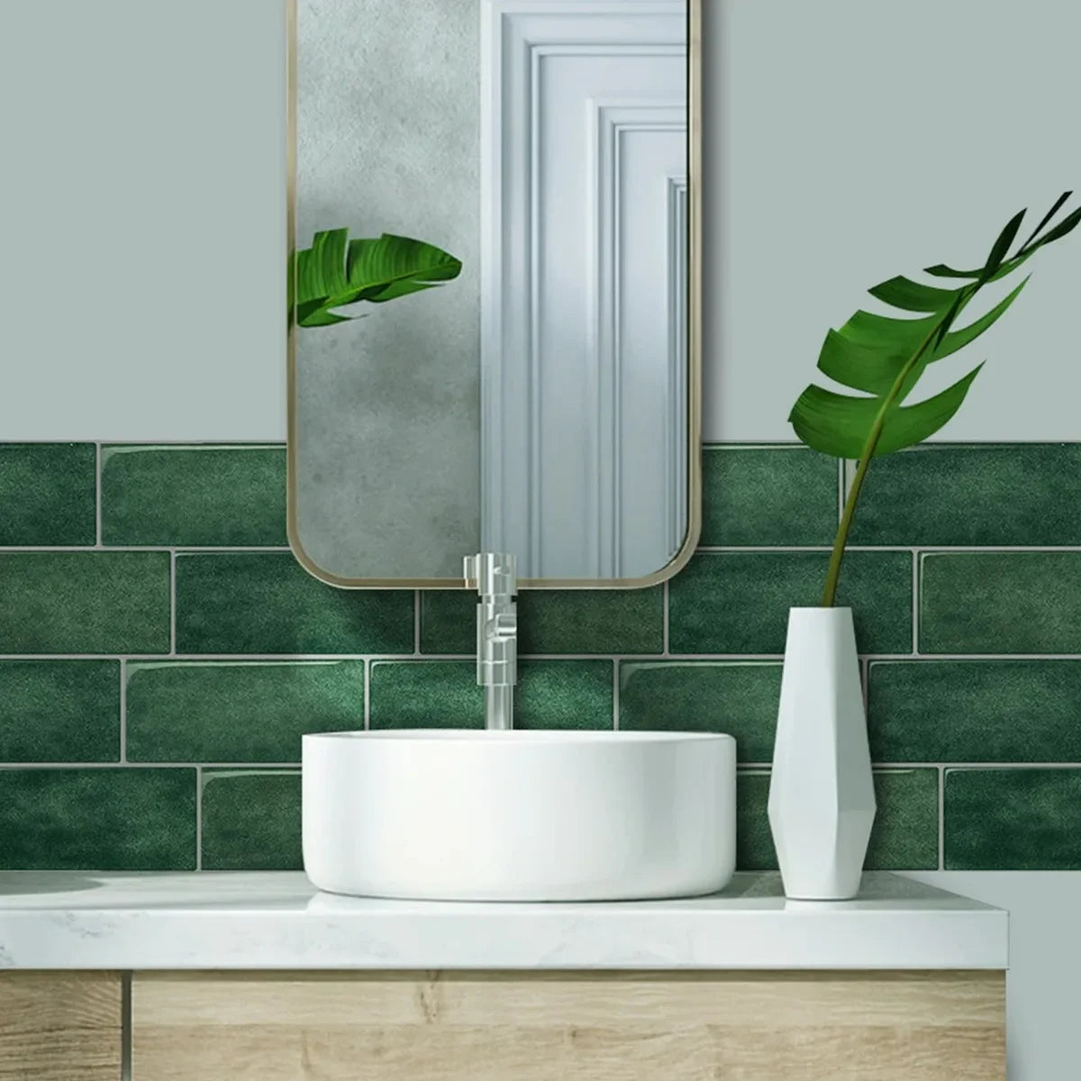 A white vessel sink is mounted on a light wood countertop against a dark green subway tile backsplash with a mirror and tropi