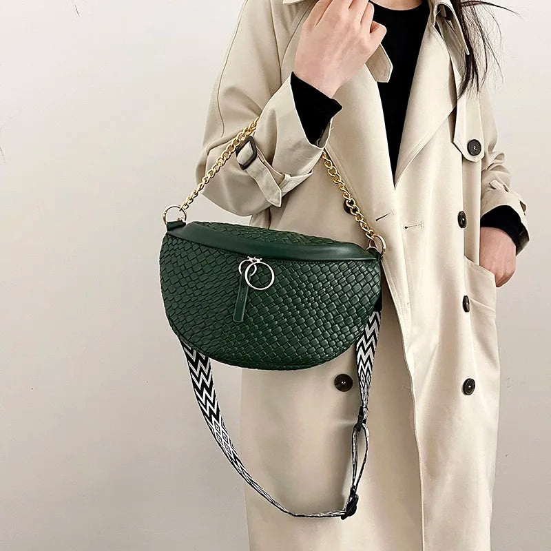 A woman holds a dark green woven pattern waist bag with gold chain strap against a light beige trench coat.