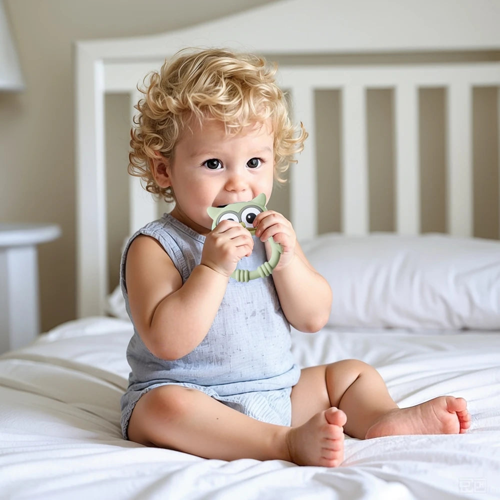 A baby is holding an owl-shaped silicone teether and rattle in a light blue outfit while lying on white bedding.