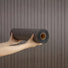 A hand holds a roll of dark brown, textured self-adhesive wallpaper with a wood grain pattern against a vertical wooden backg