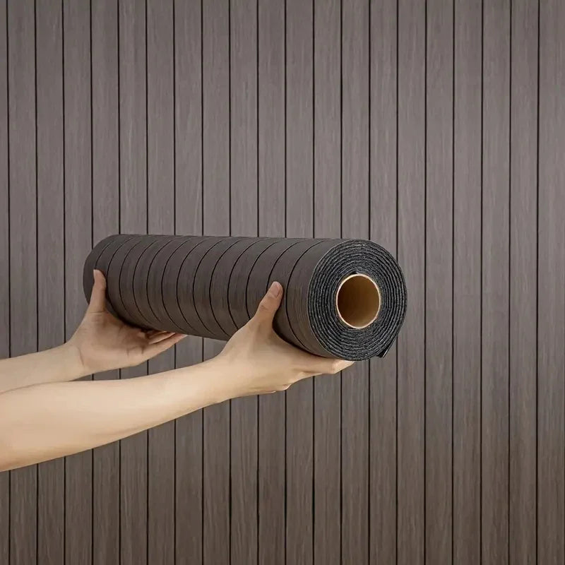 A hand holds a roll of dark brown, textured self-adhesive wallpaper with a wood grain pattern against a vertical wooden backg