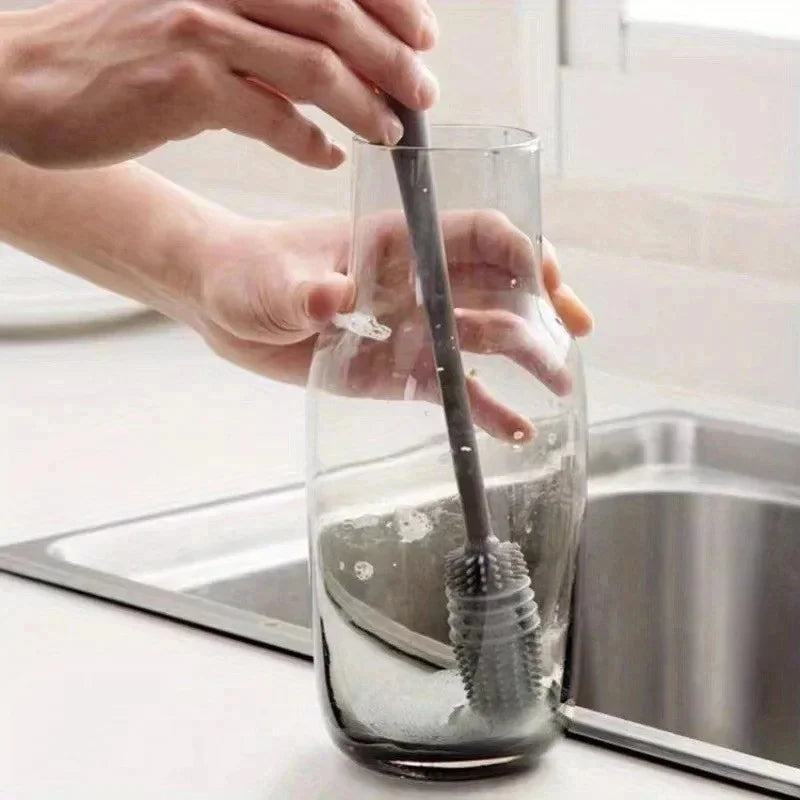 Hands are cleaning a glass bottle brush with a long handle inside a stainless steel sink.