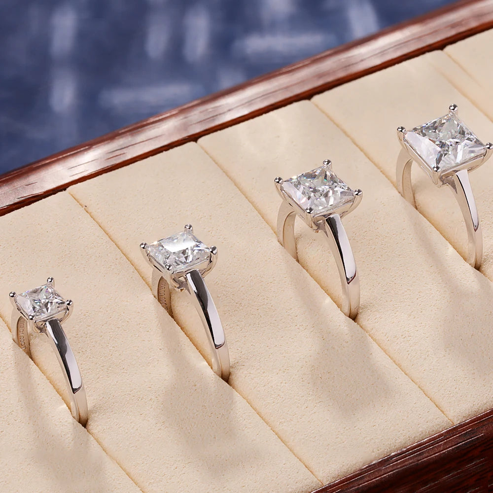 A row of princess cut moissanite solitaire rings displayed on a wooden jewelry box.