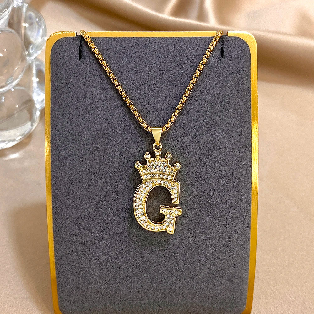 A gold letter pendant necklace featuring a crown design and a letter G, displayed on a dark gray card.