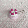 A pink and white cartoon cake clicker fidget keychain with pink frosting details resting on a pink textured surface.