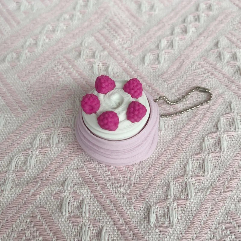 A pink and white cartoon cake clicker fidget keychain with pink frosting details resting on a pink textured surface.