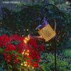 A vintage solar watering can garden light with a shepherd's hook is shown pouring glowing lights into red flowers.
