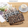 A close-up of a brown leopard print retro long wallet resting on a wooden surface next to a white cup and some greenery.