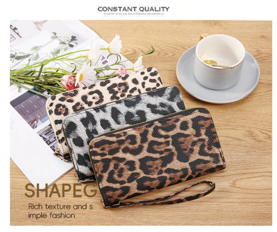 A close-up of a brown leopard print retro long wallet resting on a wooden surface next to a white cup and some greenery.