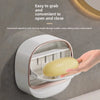 A hand is opening a wall-mounted soap holder with a lid and drainage feature.