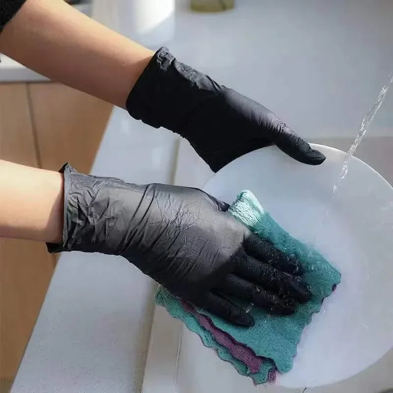 A hand wearing a black nitrile disposable glove is washing a white plate with a green cleaning cloth.