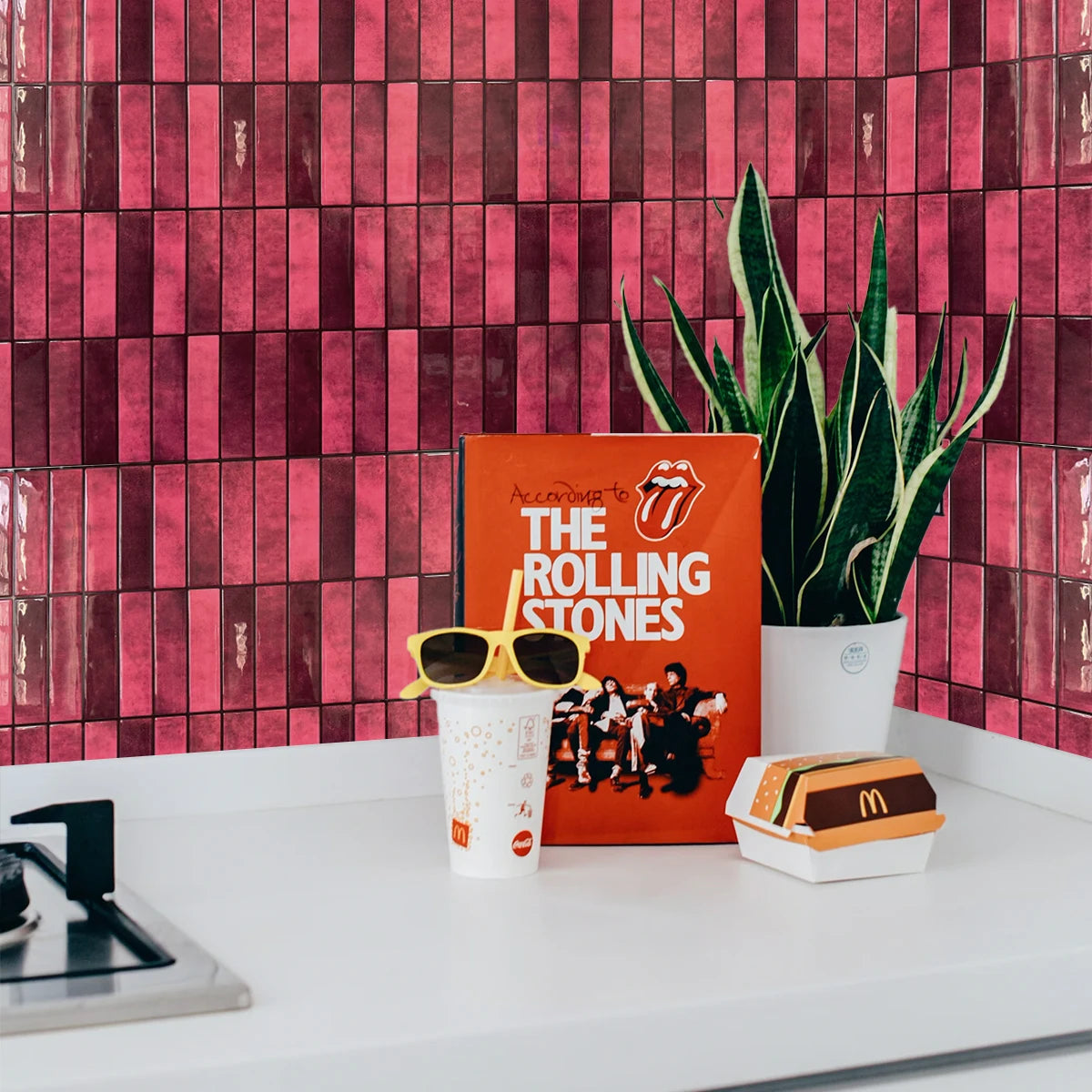 Gros plan d'un mur carrelé rose et bordeaux avec une boîte orange, des lunettes de soleil et une tasse blanche sur une surfac