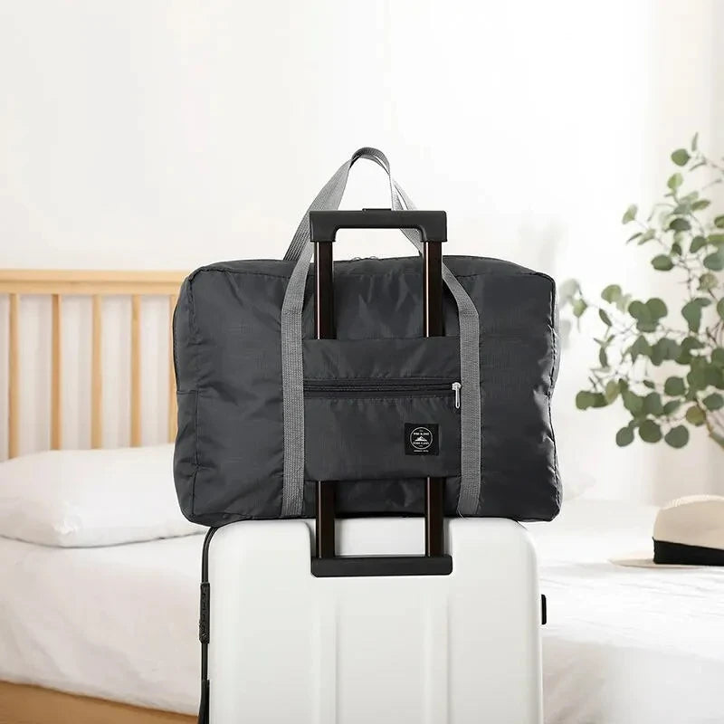 A black foldable travel duffel bag is placed on a white rolling suitcase next to a bed.