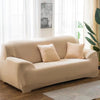 A beige elastic stretch sofa cover for a 3-seater sofa with two throw pillows, set in a bright room.