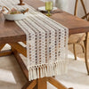 A beige macrame table runner with fringe is displayed on a wooden dining table.