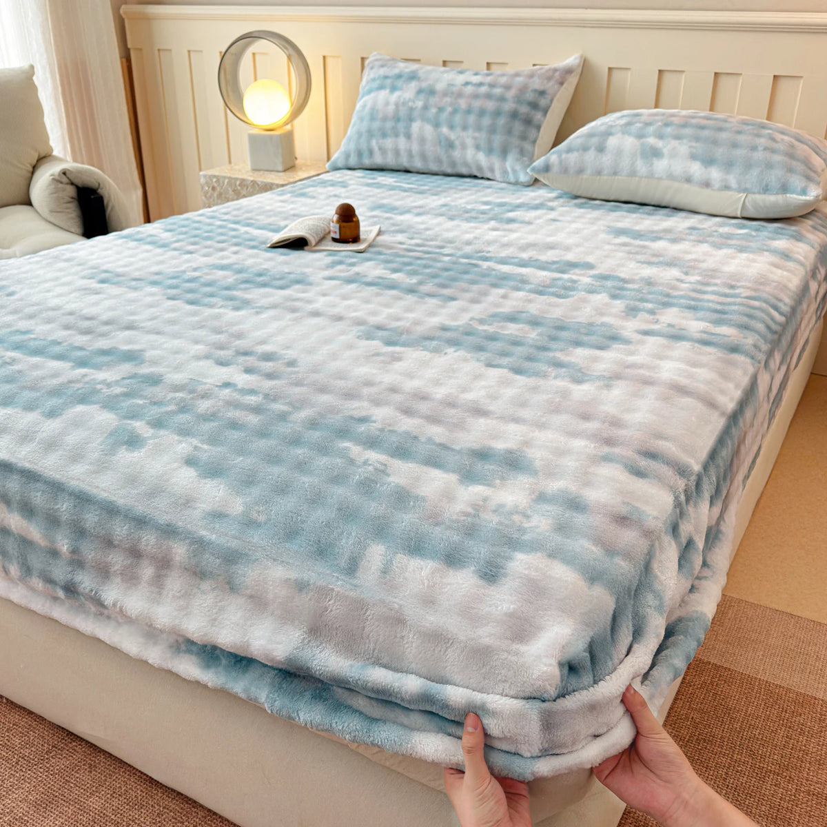 A light blue and white faux fur fitted sheet is displayed on a bed, with pillows and a lamp in the background.