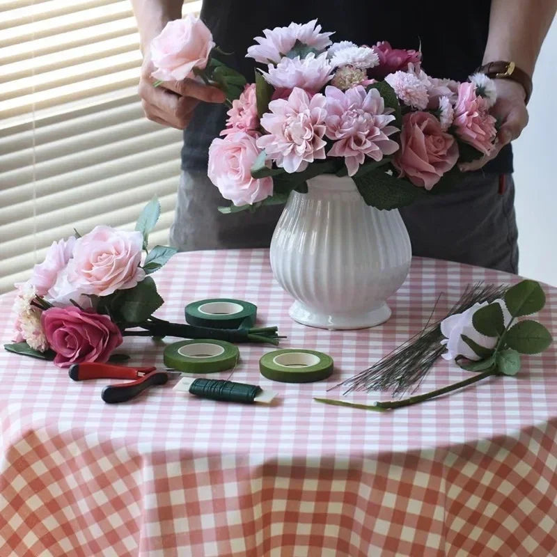 A person holds a bouquet of pink and purple flowers, with rolls of green floral tape laid out on a checkered tablecloth.