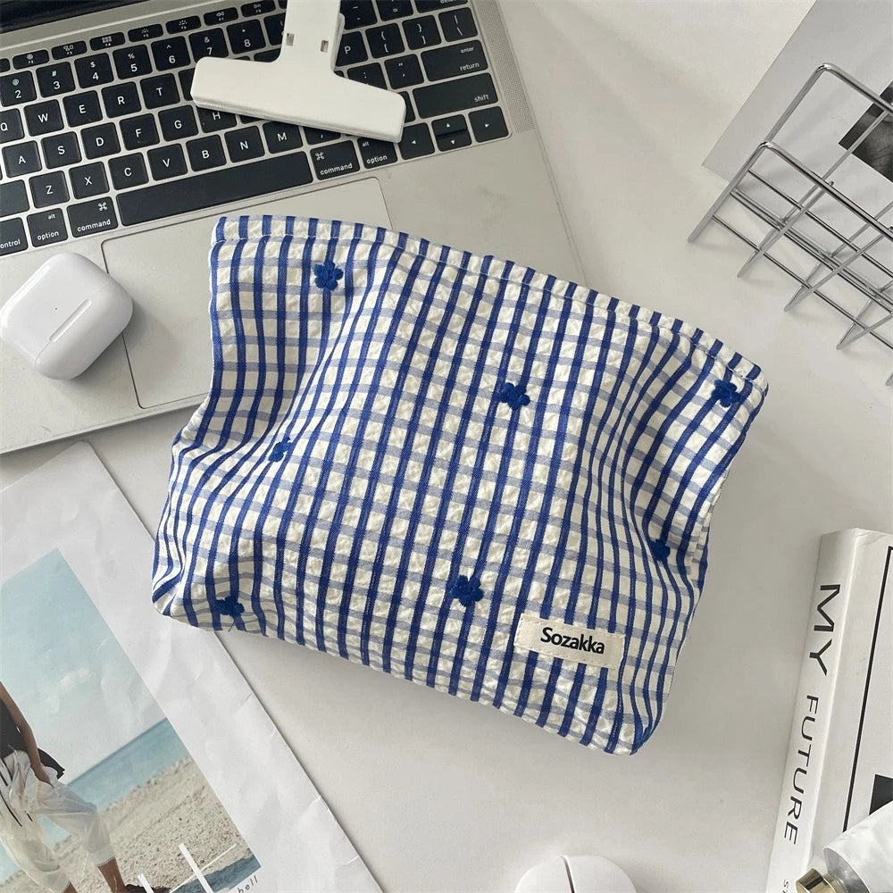 A blue and white plaid makeup bag with floral embroidery resting on a white desk next to a laptop.