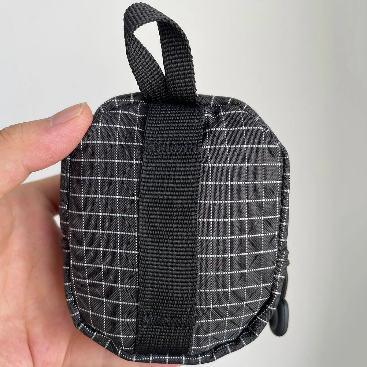 A hand holds a compact EDC utility pouch made of black and white plaid fabric with a black strap.