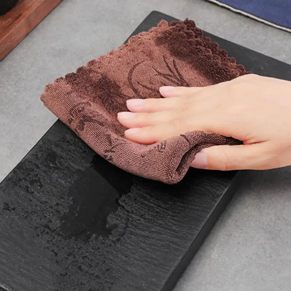 Eine Hand hält ein dunkelbraunes, strukturiertes saugfähiges Handtuch auf einer schwarzen Schieferoberfläche.