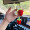 A hand holds a handmade crochet strawberry pendant with white flowers, hanging in front of a car interior.
