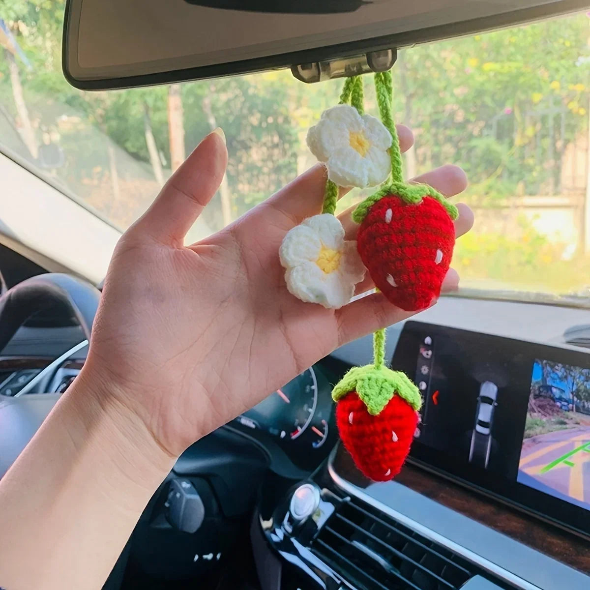 A hand holds a handmade crochet strawberry pendant with white flowers, hanging in front of a car interior.