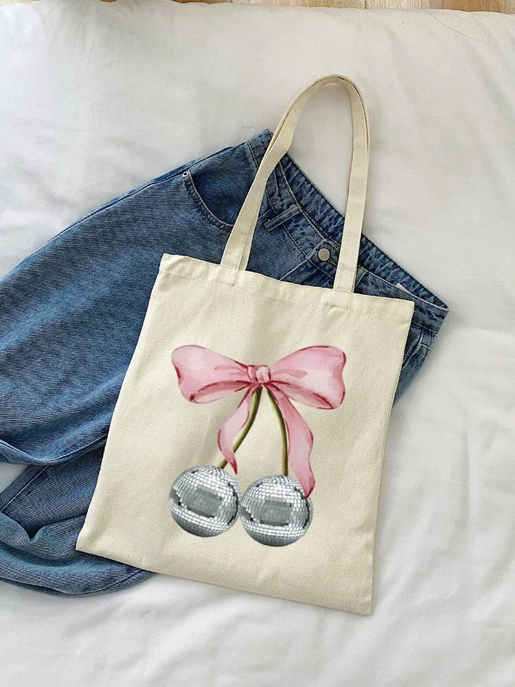 A canvas tote bag with a pink bow and two silver spherical charms resting on a white sheet.
