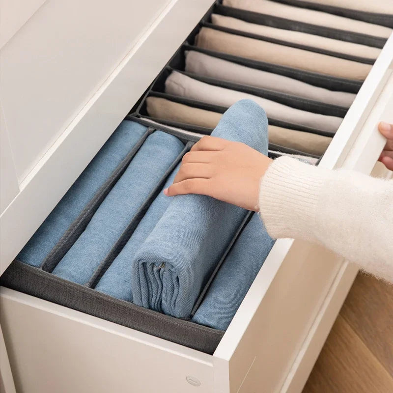 A hand is placing folded blue fabric items into a light gray drawer organizer for clothing.