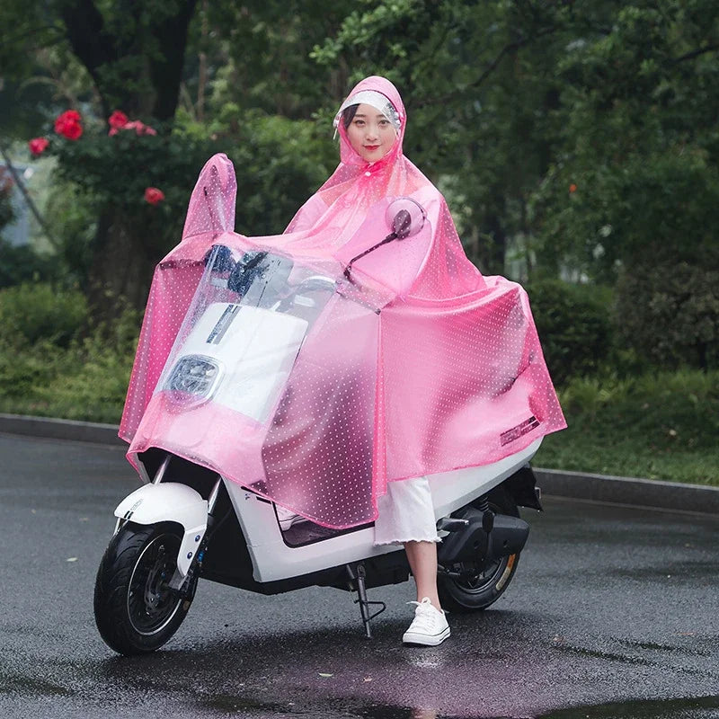 A woman wearing a pink transparent waterproof motorcycle rain poncho with a hood riding a white scooter on a wet road.